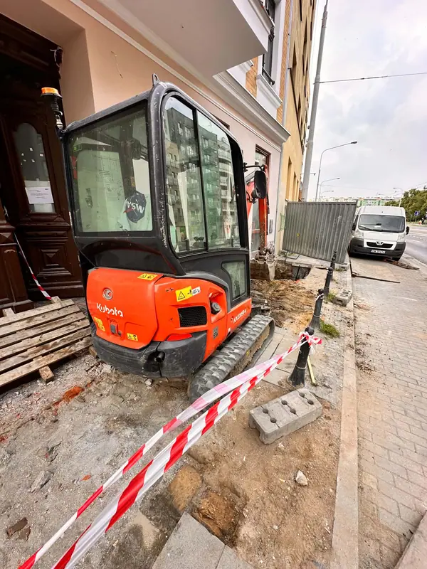 Minikoparka pracująca w wąskim przejściu między ścianą budynku a chodnikiem, ogrodzona taśmą ostrzegawczą.