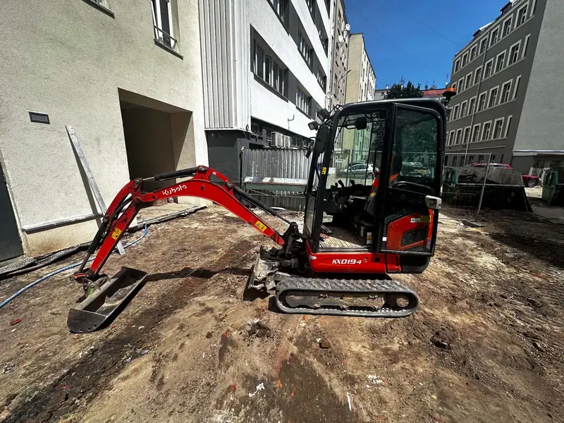 Minikoparka Kubota KX019-4 podczas kopania wykopu na terenie miejskim, między budynkami.